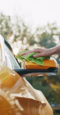 Bir kişi yeşil bir bezle sarı bir arabanın yan aynasını temizliyor, araba bakımını ve bakımını dikey bir şekilde gösteriyor..