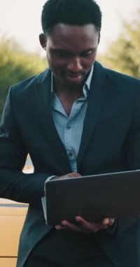 Takım elbiseli bir siyahi işadamının dikey çekimi. Açık havada dizüstü bilgisayarı üzerinde çalışıyor. Modern iş ve teknolojiyi gözler önüne seriyor..