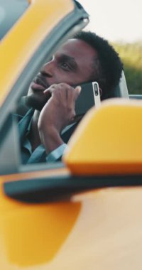 Takım elbiseli gülümseyen siyah bir adam, üstü açık sarı bir arabaya binerken telefonuyla konuşur. Dikey video.