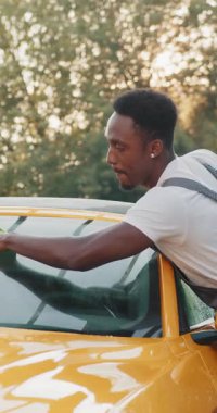 Sarı bir arabanın ön camını, yeşil arka planda mikrofiber kumaşla temizleyen bir adam görülüyor..