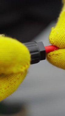 Sarı eldiven takmış siyah bir objeyi, muhtemelen bir alet ya da bileşeni manipüle eden ellerin yakın çekimi. Arka plan bulanık, harekete odaklanmış durumda..