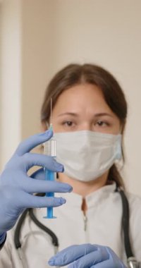 Maske ve eldiven takan bir tıp uzmanı enjeksiyon için şırınga hazırlar, sağlık hizmetleri ve aşı kavramlarını vurgular.