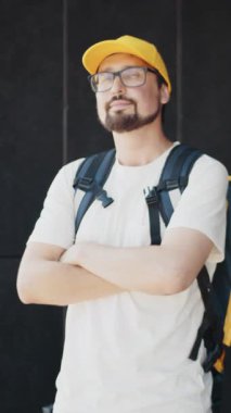 Sakallı, gözlüklü ve sarı şapkalı bir adam kollarını kavuşturup sırt çantası takıyor..