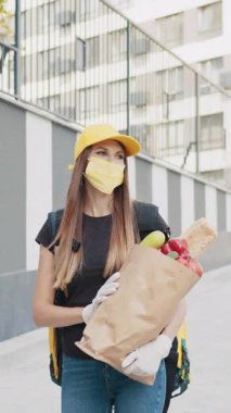 Sarı şapkalı ve maskeli genç bir kadın, muhtemelen sağlık sorunları sırasında market poşeti getiriyor. Eldiven ve teslimat çantası takıyor..