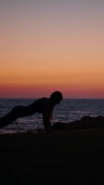 Bir kadın, okyanusun üzerinde canlı bir günbatımına karşı siluet şeklinde tahta poz veriyor. Dikey video.