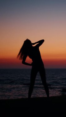 Bir kadın silueti okyanusun üzerinde canlı bir günbatımına karşı çekilir, sakin ve düşünceli bir sahne yaratır..