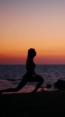 Bir kadın gün batımında plajda yoga yapıyor, renkli gökyüzüne karşı siluet. Bu dikey video dingin anı yakalar.