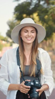 Şapka ve beyaz gömlek giyen bir fotoğrafçı dışarıda profesyonel bir kamerayla fotoğraf çekmeye odaklanır. Dikey video.