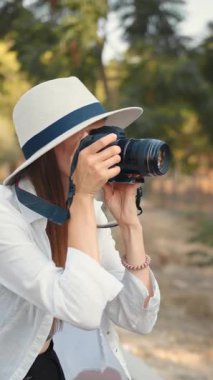 Beyaz şapkalı ve gömlekli güzel bir kadın dışarıda profesyonel bir kamerayla fotoğraf çekerken gülümsüyor..