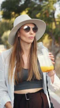 Güneşli bir günde güneş gözlüğü ve şapka takan bir kadın ferahlatıcı bir portakal suyu içmekten hoşlanır..