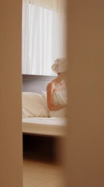 Havluya sarılmış bir kadın, dizüstü bilgisayarıyla yatakta otururken çalışıyor. Bu dikey çekim bir rahatlama ve üretkenlik anı yakalar..