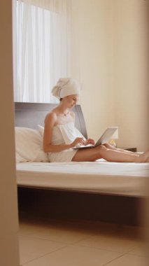 Havluya sarılmış bir kadın, dizüstü bilgisayarıyla yatakta otururken çalışıyor. Dikey video.