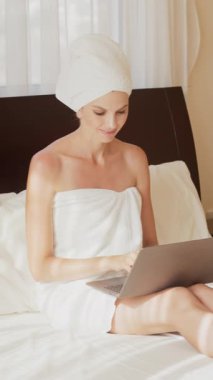 Kafasına havlu takmış bir kadın yatakta oturur ve dizüstü bilgisayarla çalışır. Dikey video.