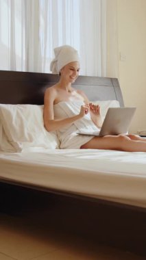 Havluya sarılmış bir kadın, dizüstü bilgisayar kullanırken yatakta oturup gülümsüyor. Dikey video.