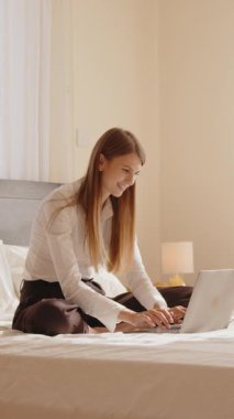 Genç bir kadın yatakta bacak bacak üstüne atmış bir şekilde oturuyor ve gülümseyerek dizüstü bilgisayarla çalışıyor. Rahat ve iyi aydınlatılmış bir yatak odası ortamında odaklanmış görünüyor..
