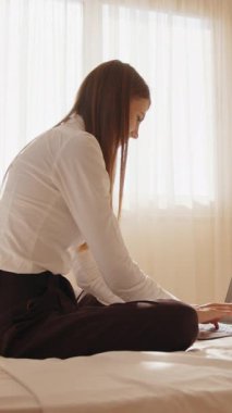 Bir kadın, dizüstü bilgisayarla güneşli bir yatak odasında bacak bacak üstüne atarak oturuyor. Bu dikey çekim uzak bir çalışmayı yakalar..