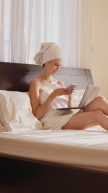 Havluya sarılmış bir kadın yatakta dinlenirken telefonunu ve laptopunu kullanıyor. Dikey video.
