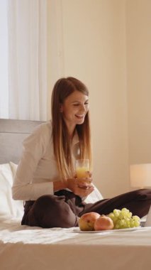 Bir kadın yatakta oturmuş, bir bardak meyve suyu ve bir tabak meyvenin tadını çıkarıyor, rahatlama ve refah sahnesi yaratıyor..