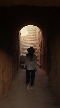 Bir kadın kadim, kemerli bir yapı içinde merdivenlerden yükselir, tepede güneş ışığıyla yıkanır. Dikey video.