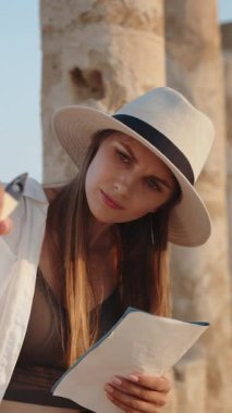 Eski sütunların önünde dururken, şapka takan bir kadın bir kağıt bloknotun üzerine resim çiziyor. Dikey video.