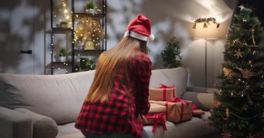 Noel Baba şapkası takan bir kadın, Noel günü etrafı şenlikli süslemeler ve Noel ağacı ile çevrili bir hediyeyi açar. Noel Baba şapkası takan bir kadın Noel günü hediye kutusunu açar.