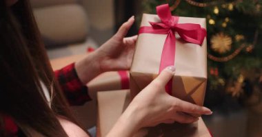 Bir insan elinde kırmızı kurdeleli bir hediyeyle bir Noel ağacının önünde duruyor. Bir insan süslenmiş bir Noel ağacının yanında paketlenmiş hediyeler tutuyor ve bayram sahnesi yaratıyor.