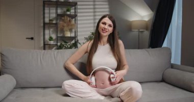 Hamile kadın kanepeye oturup karnına kulaklık takarak doğmamış bebeğine müzik çalıyor. Hamile Kadın Müzik Dinliyor