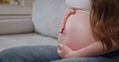 Kadınlar koltukta otururken karnını nazikçe kucaklıyor. Beklenti ve annelik sevgisini gösteriyor. Hamile Göbeği Kapat