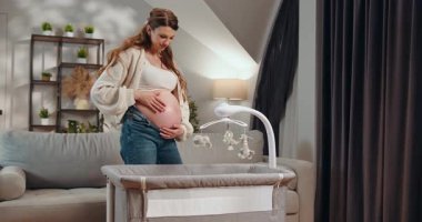 Hamile kadın rahat bir odada cep telefonuyla bebek karyolasının yanında dururken karnına dokunuyor. Bebek Beşiğine Hamile Kadın