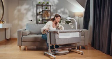 Kadın yatağının yanında dinleniyor. Cep telefonu var. Rahat oturma odası ortamında bitkin hissediyor. Yorgun Anne Beşiğin Yanında