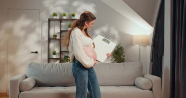 Hamile bir kadın oturma odasında tartıyı tutuyor, hamilelik süresince kilo alımını izliyor, mutlu ve sağlıklı hissediyor. Ölçekli Hamile Kadın