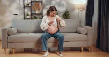 Bekleyen anne kanepeye oturur, geniş karnını evde düşünceli bir ifadeyle sarmalar. Hamile Kadın Koltukta Göbeğe Dokunuyor