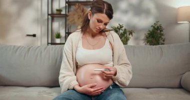 Hamile bir kadın kanepeye oturmuş hamilelik testi setini tutuyor ve düşünceli bir ifadeyle bakıyor. Hamile Kadın Hamilelik Testi