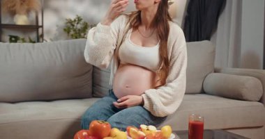 Hamile kadın kanepede karnını tutarak oturur ve küçük aperatifler yer, kendini rahat ve rahat hisseder. Hamile Kadın Evde Atıştırmalık Yiyiyor