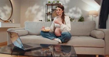 Hamile kadın kanepede bağdaş kurup yüzünde endişeli bir ifadeyle elinde patlamış mısırla oturuyor. Hamile kadın koltukta patlamış mısır yiyor.