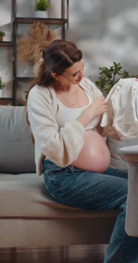 Hamile kadın kanepeye oturur, kucağında bebek kıyafetleri, yeni doğan bebeğin gelişini nazikçe gülümseyerek bekler.