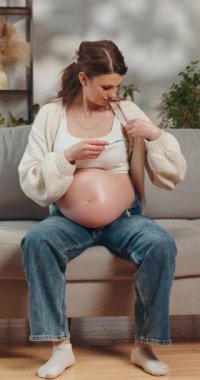 Bekleyen anne, kanepede rahat bir şekilde otururken karnını kocaman açar. Nazik bir ifadeyle aşağı bakar, kapalı bir yerde.