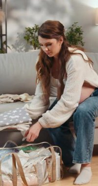 Uzun kahverengi saçlı, krem rengi kazak giyen ve kot pantolon giyen genç bir kadın bir koltukta oturur ve kucağındaki bir şeye bakar.