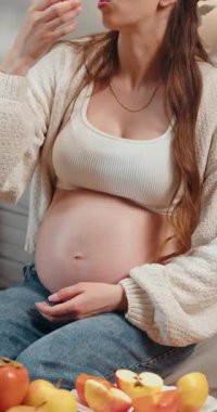 Hamile bir kadın karnını tutuyor ve küçük sarı atıştırmalıklar yiyor. Mutlu ve rahat hissediyor.