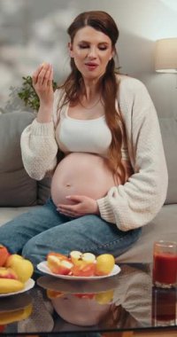 Hamile kadın gündelik kıyafetlerle kanepede oturup küçük sarı yemek yiyerek karnını gösteriyor.