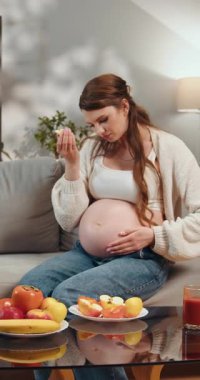 Kanepede oturan hamile bir kadın, elinde bir parça meyve, kahve masasında çeşitli meyveler, iç mekan.