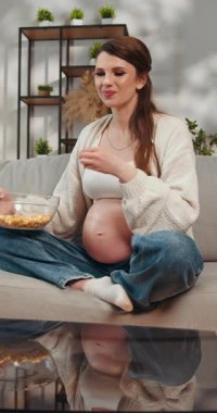 Kanepede oturup kaseden patlamış mısır yiyen hamile bir kadın.