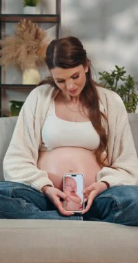 Hamile kadın kanepeye oturmuş elinde telefonla bebek ayaklarının ultrason görüntülerini gösteriyor, anneliği ve yeni hayatı tahmin ediyor.