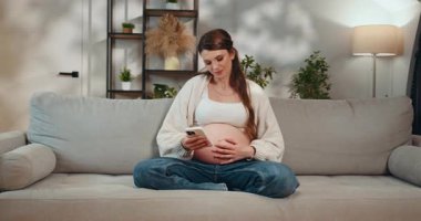 Bekleyen anne kanepede bağdaş kurarak oturur, telefonu tutar ve karnına dokunur, rahat bir iç mekan ayarlar. Kanepede akıllı telefon kullanan hamile kadın.