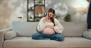 Hamile bir kadın koltukta oturmuş, karnını tutarken telefonla konuşuyor, arka planda bitki ve raflarla iç mekanlar hazırlıyor. Telefonda Konuşan Hamile Kadın