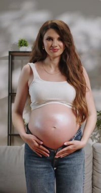 Uzun kahverengi saçlı, beyaz kolsuz bluzlu ve kot pantolonlu genç bir kadın hamile karnını nazikçe gülümsüyor.