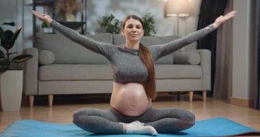 Atletik giysili hamile bir kadın yoga minderinde bacak bacak üstüne atmış, elleri dua pozunda birbirine bastırılmış, oturma odasında farkındalık ve rahatlama pratiği yapıyor. Hamile Kadın Evde Meditasyon Yapıyor