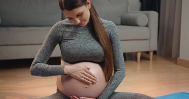 Gri spor elbiseli hamile bir kadın yere oturup karnına hafifçe dokunur ve rahat oturma odasında bebek hareketlerini hisseder. Hamile Kadın Karnına Dokunuyor