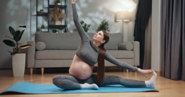 Gri aktif giysili hamile kadın oturma odasında yoga minderinde yan esneme hareketleri yapıyor ve hamilelik süresince sağlığını arttırıyor. Hamile Kadın Evinde Esniyor