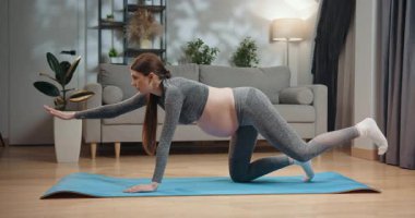 Spor giyim bölümünde hamile bir kadın oturma odasında yoga minderinde kuş-köpek pozu veriyor, hamilelik süresince çekirdek dayanıklılığına ve dengesine odaklanıyor. Hamile Kadın Evde Yoga Yapıyor